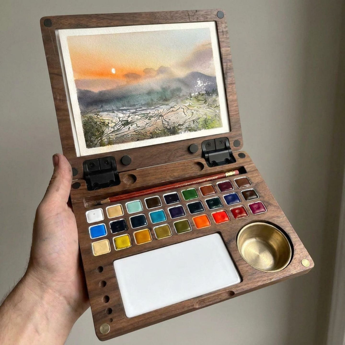 Hand holding the open ArtMate Master Edition walnut watercolor box, a finished sunset rice-terraces painting clipped to the lid above the 27-pan palette, mixing tile and brass water pot