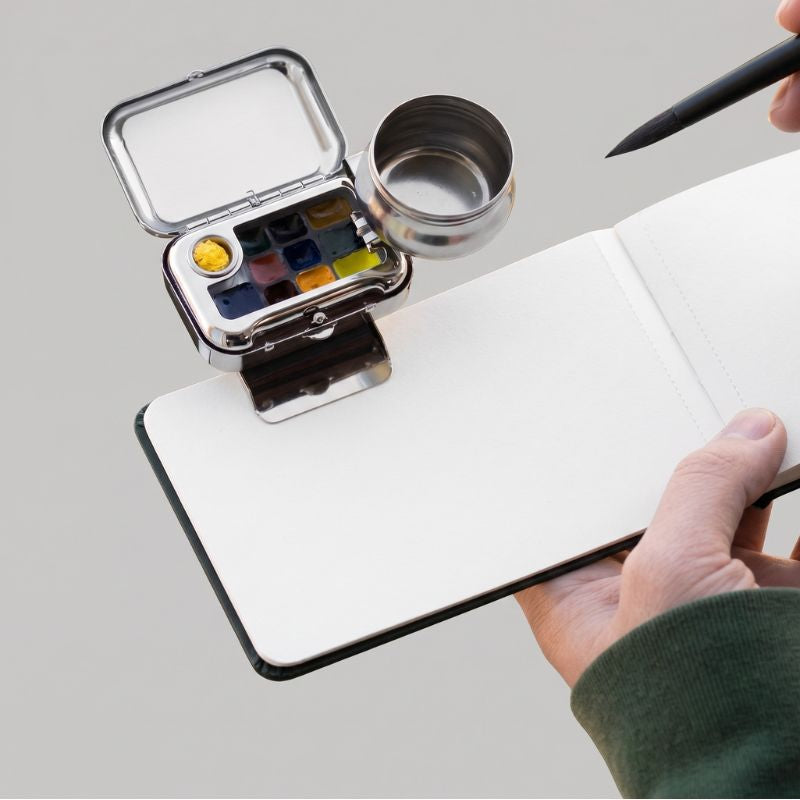 Artist painting with an open ArtMate travel watercolor kit and brush over a sketchbook