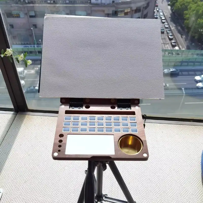 Walnut painting kit on a tripod by a window with a city view and blank watercolor paper clipped