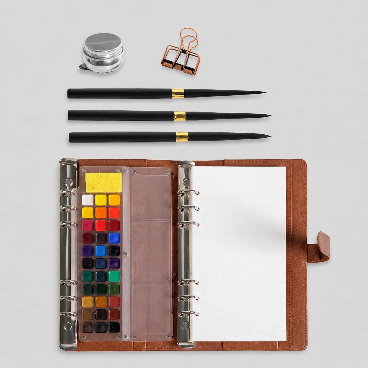 Flat lay of the open Voyager kit showing the 33-color magnetic palette, absorbent sponge and mixing area, with three travel brushes, a water pot and a binder clip