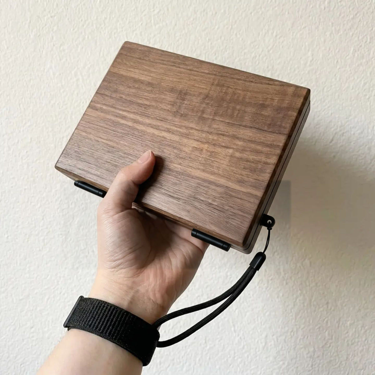 Hand holding the closed walnut Master Edition box from above, showing the wood-grain lid and black wrist strap