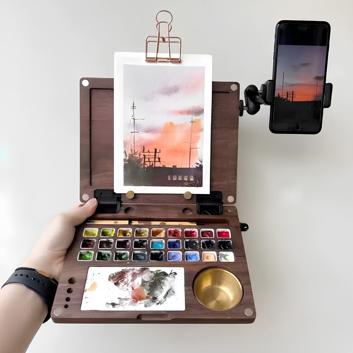 Open walnut Master Edition station held in hand with a sunset painting clipped to the lid, a 27-pan palette and brass water pot, and a phone mounted on the adjustable arm as a reference