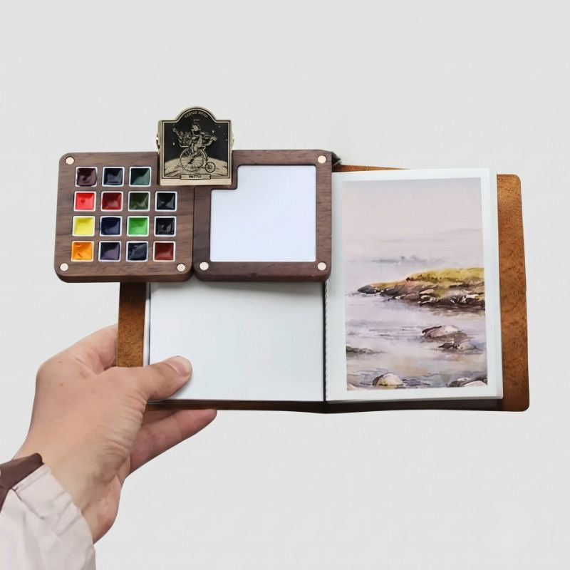 Open ArtMate Adventure Artist Kit held by a rocky shore, 15-color palette and a seascape painting in the sketchbook
